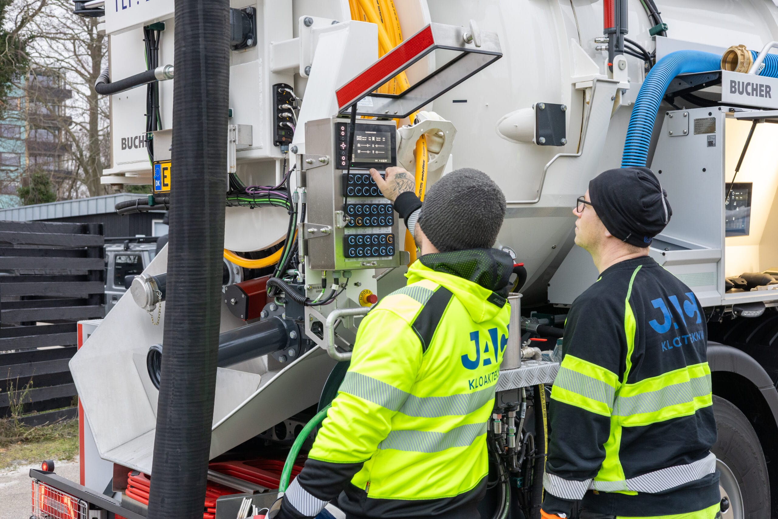 To medarbejdere fra JAC Kloakservice står bag en slamsuger og betjener kontrolpanelet på bilen under arbejde i et boligområde. På deres jakker står "JAC KLOAKTEKNIK" mens de justerer indstillinger på udstyret for at udføre kloakservice effektivt.