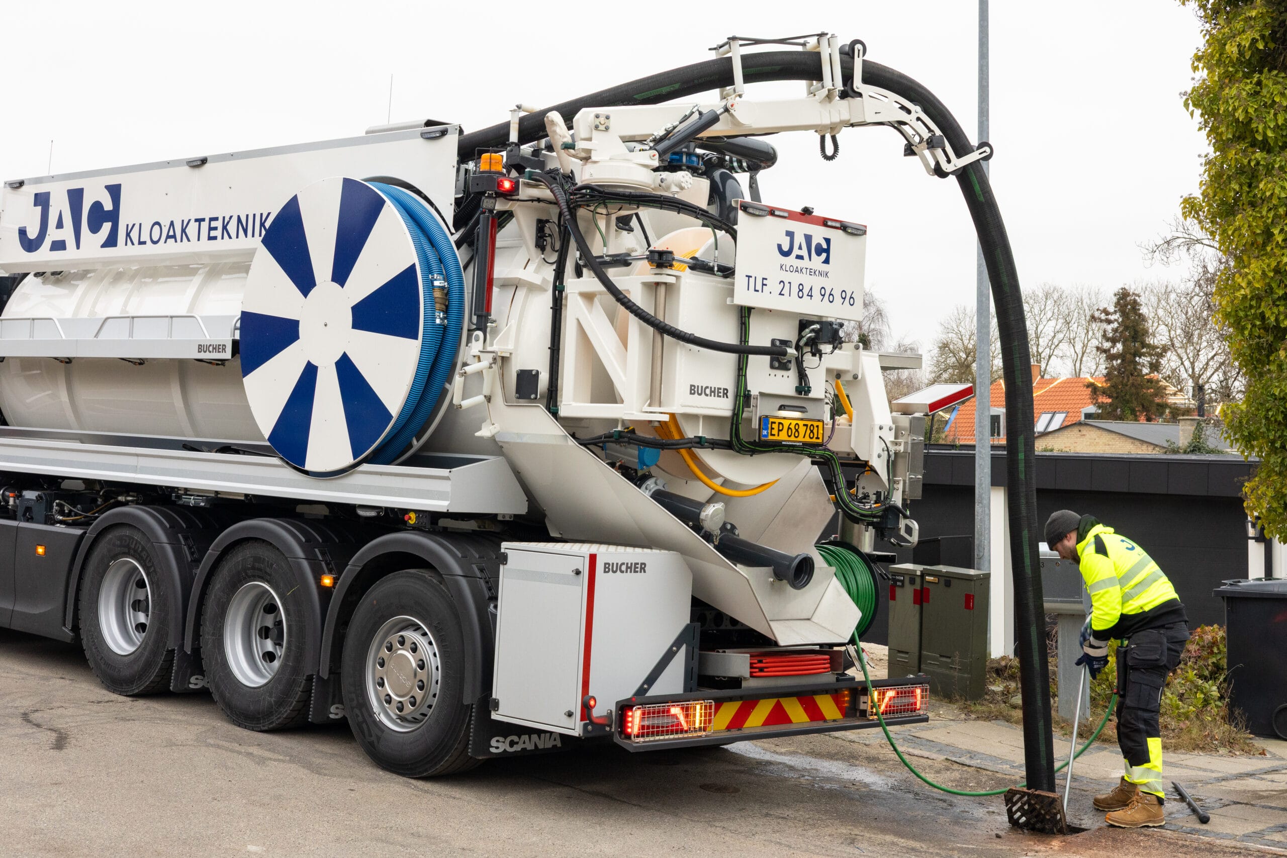En stor slamsuger fra "JAC KLOAKTEKNIK" holder parkeret ved vejkanten mens en medarbejder udfører kloakservice med en lang sort sugeslange ned i en brønd. På siden af lastbilen ses teksten "JAC KLOAKTEKNIK" og "TLF. 21 84 96 96" samt mærket "BUCHER". Billedet viser professionel JAC kloakservice i et boligområde med fokus på udstyr og arbejdsproces.