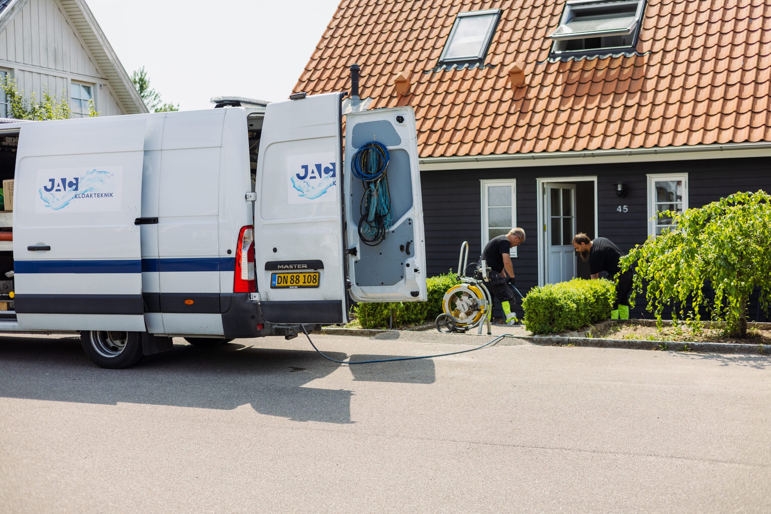 To kloakteknikere fra JAC Kloakteknik arbejder foran et hus med udstyr koblet til en hvid servicevogn med åbne døre.