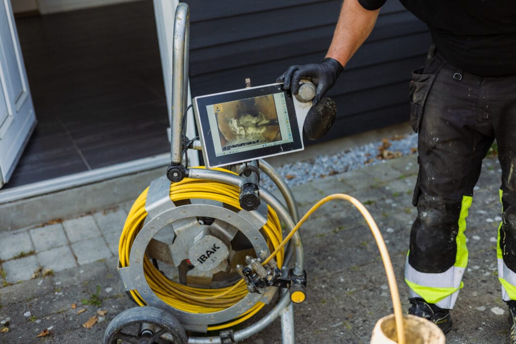TV-inspektionsudstyr med gul kabelrulle og skærm, der viser livebillede fra kloakrør, betjenes af tekniker i arbejdstøj og handsker.