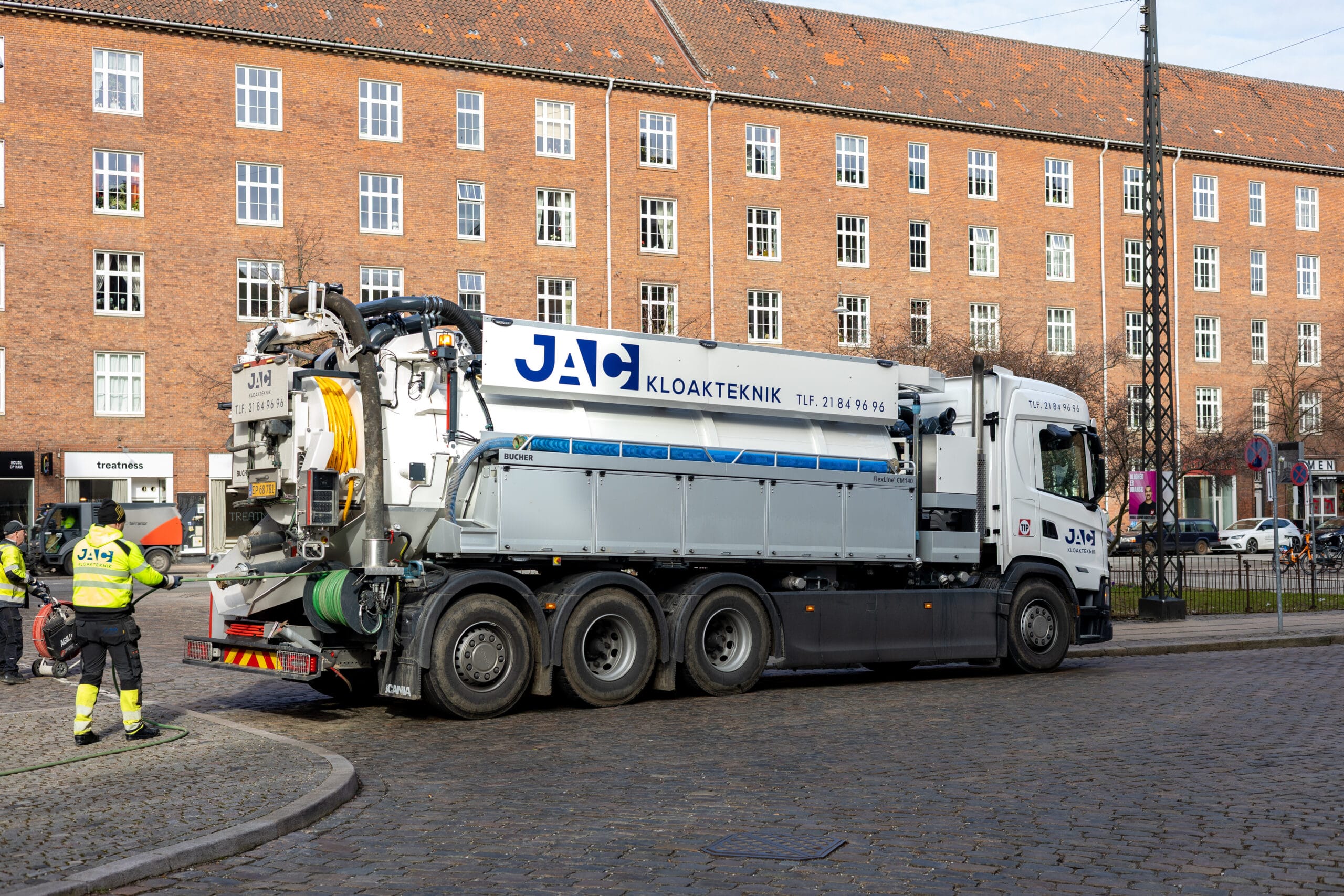 Slamsuger fra JAC kloakteknik holder på en brostensgade i København mens to medarbejdere arbejder med slanger og udstyr til faldstammerens. Lastbilen har teksten "JAC KLOAKTEKNIK" og er placeret foran en stor murstensbygning i et bymiljø.