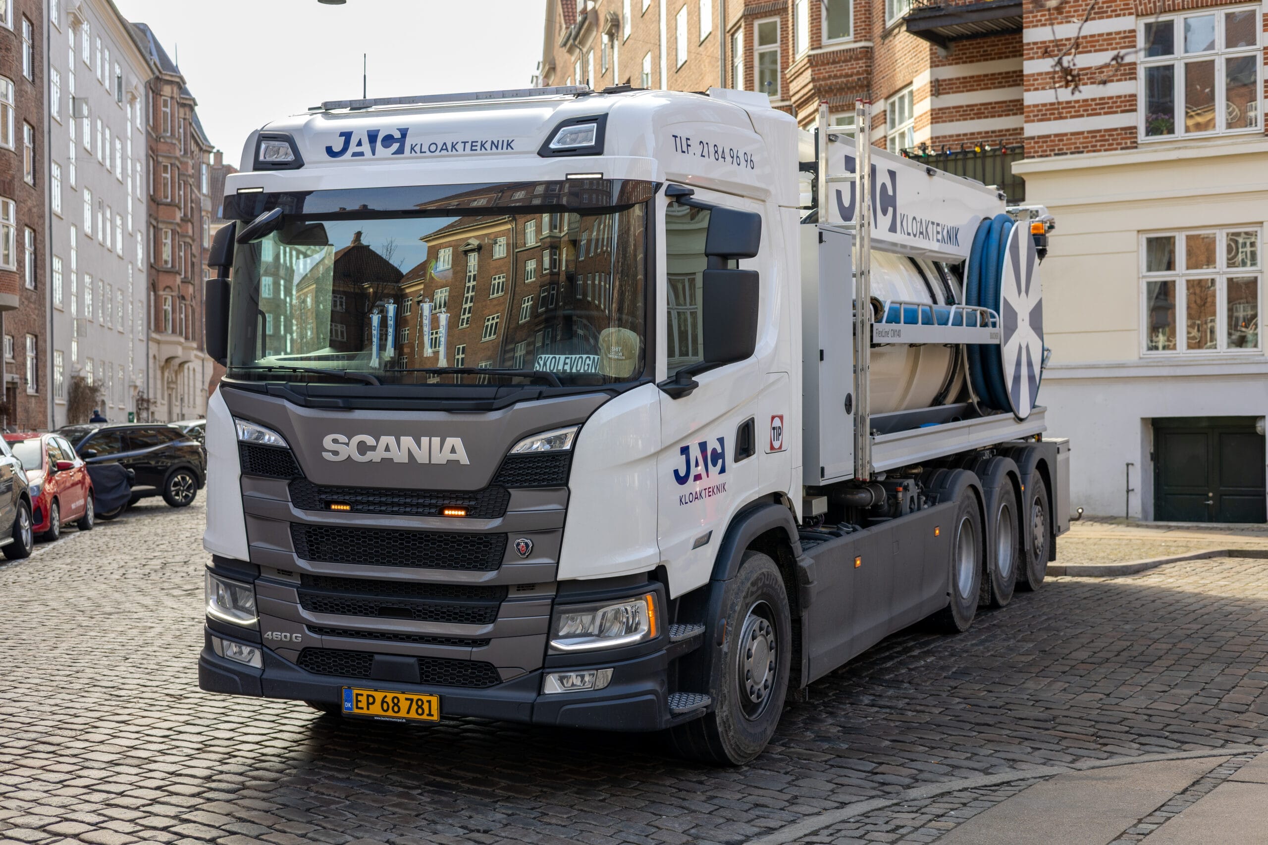 Slamsuger fra JAC kloakteknik holder parkeret på en brostensgade i København med teksten "JAC KLOAKTEKNIK" synlig på siden som en del af faldstammerens arbejde. Lastbilen er omgivet af klassiske murstensbygninger og parkerede biler i et bymiljø.