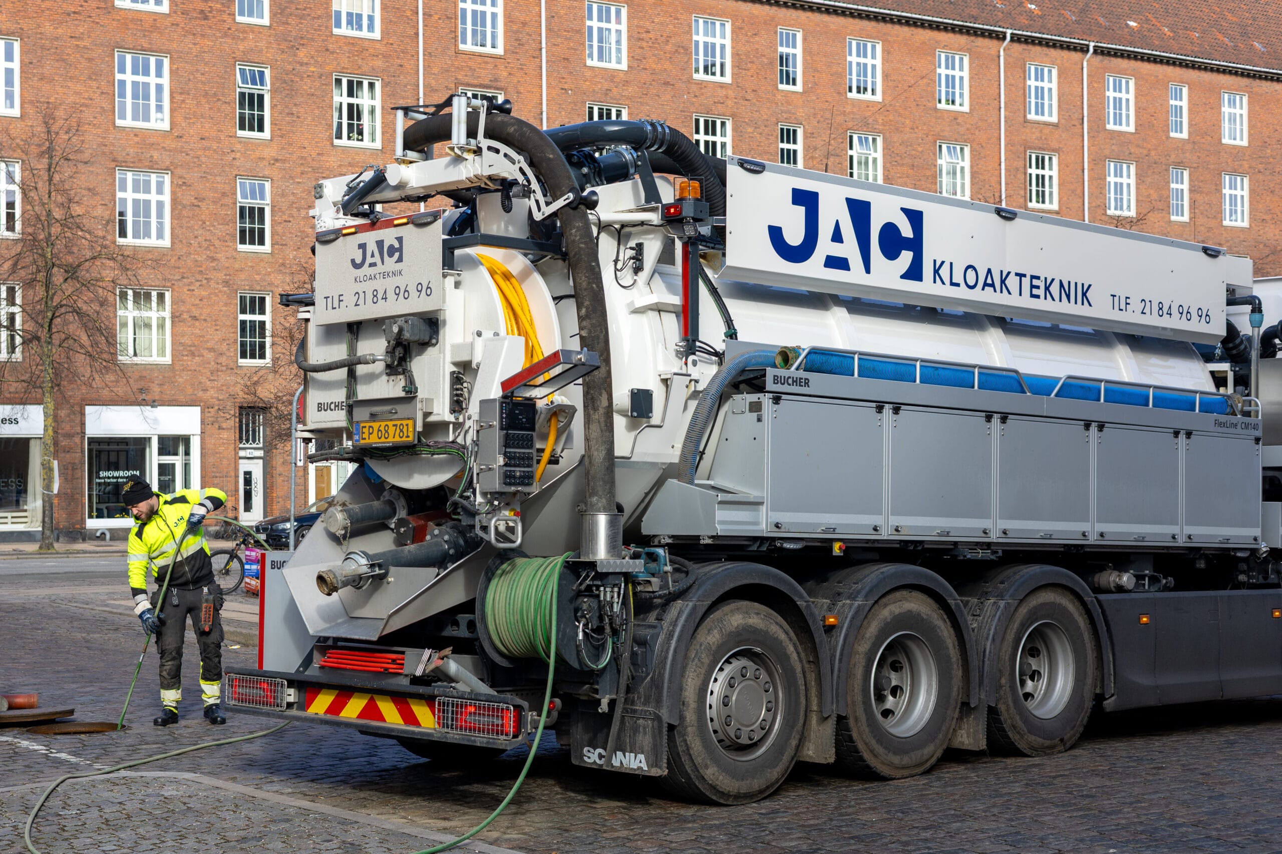 Medarbejder fra JAC kloakteknik arbejder bag en slamsuger med teksten "JAC KLOAKTEKNIK" og "Tlf. 2184 96 96" på en brostensgade i København under faldstammerens. Han håndterer en slange tilsluttet lastbilen foran en stor murstensbygning i byen.
