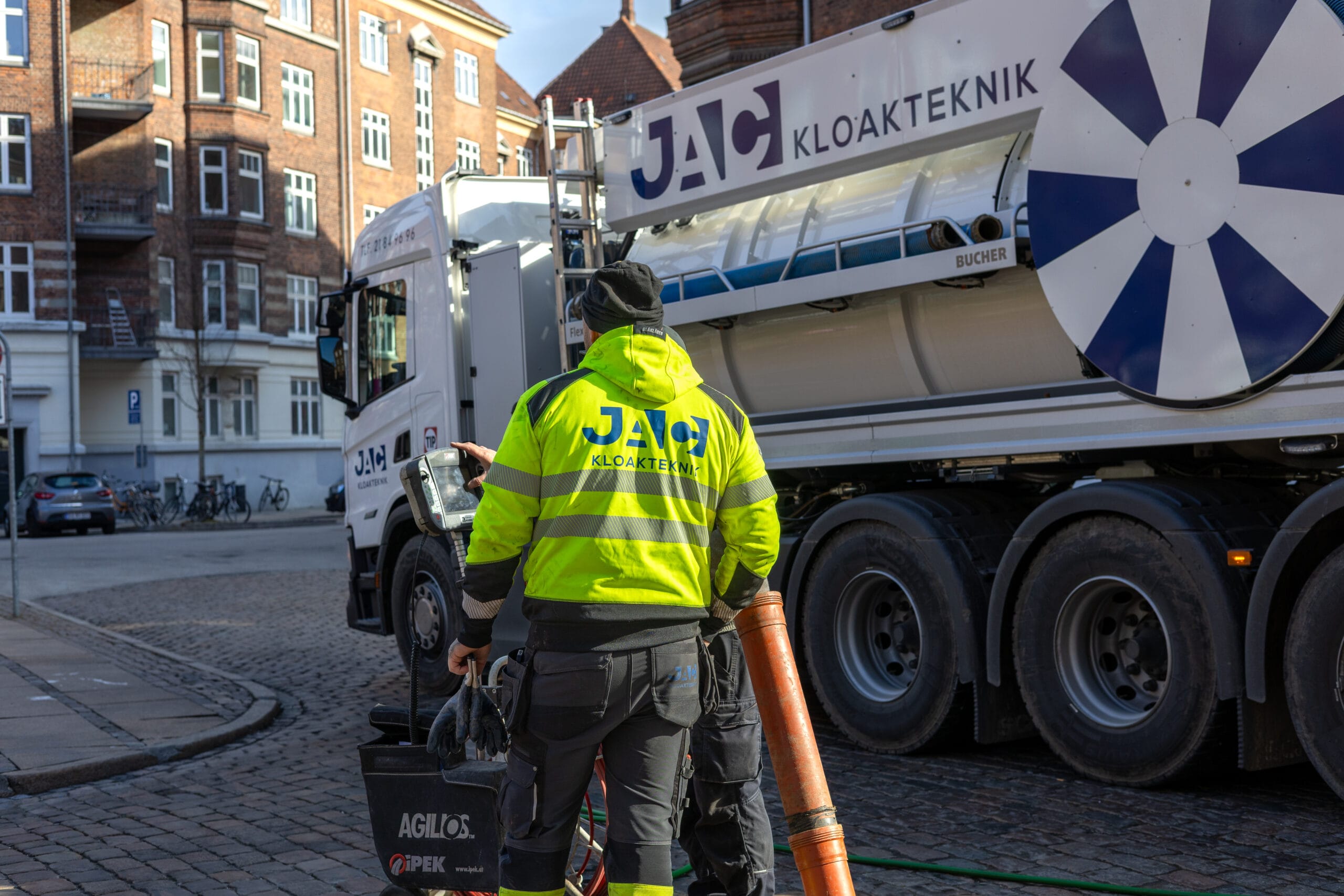 Medarbejder fra JAC kloakteknik står på en brostensgade i København foran en stor slamsuger med teksten "JAC KLOAKTEKNIK" og arbejder med udstyr til faldstammerens. Han bærer en gul refleksjakke med "JAC KLOAKTEKNIK" på ryggen og holder en slange ved siden af lastbilen mens ældre murstensbygninger ses i baggrunden.