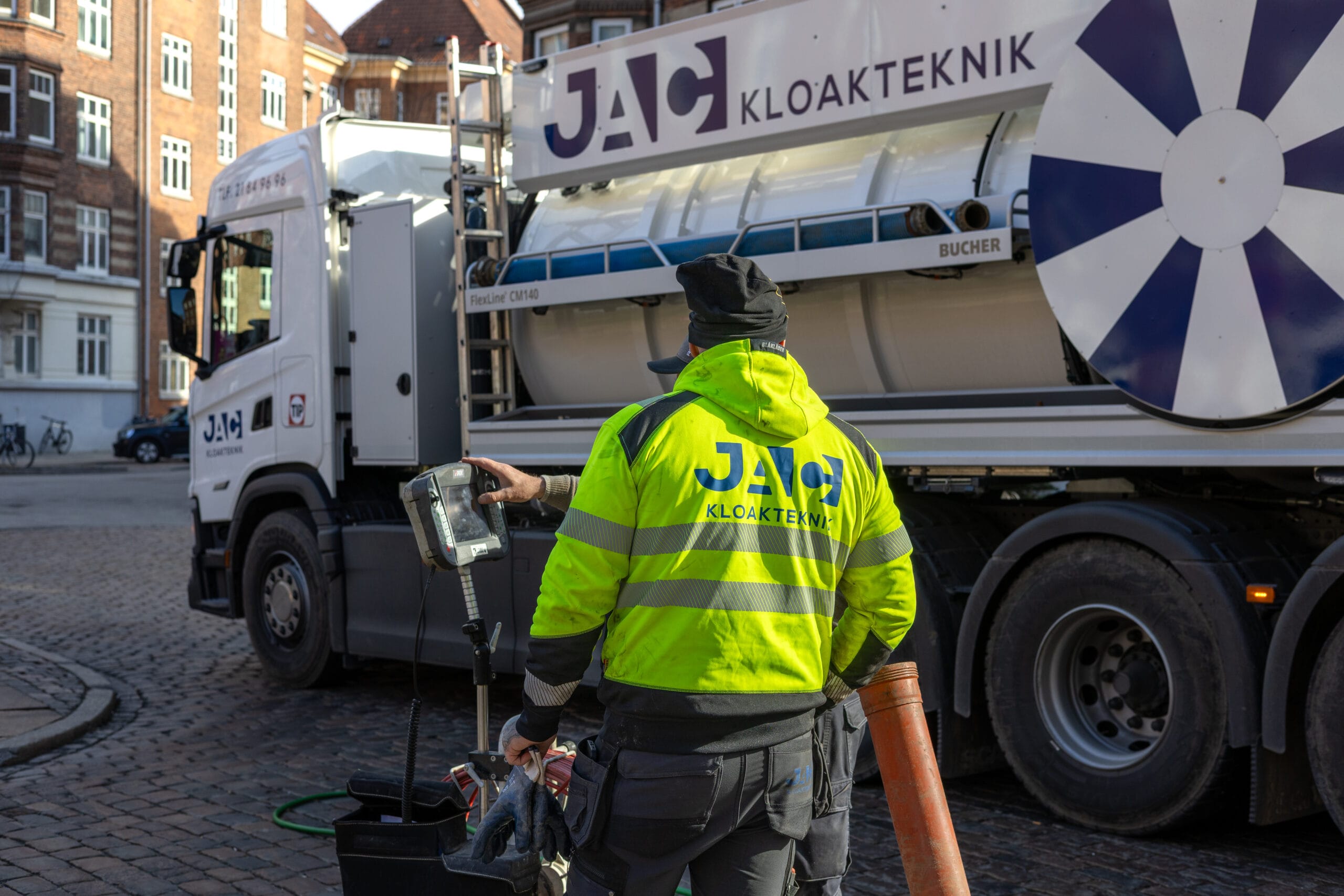 Medarbejder fra JAC kloakteknik står på en brostensbelagt gade i København ved siden af en slamsuger med teksten "JAC KLOAKTEKNIK" og arbejder med udstyr til faldstammerens. Han bærer en gul refleksjakke med "JAC KLOAKTEKNIK" på ryggen og holder en styreenhed samt en slange foran ældre murstensbygninger.