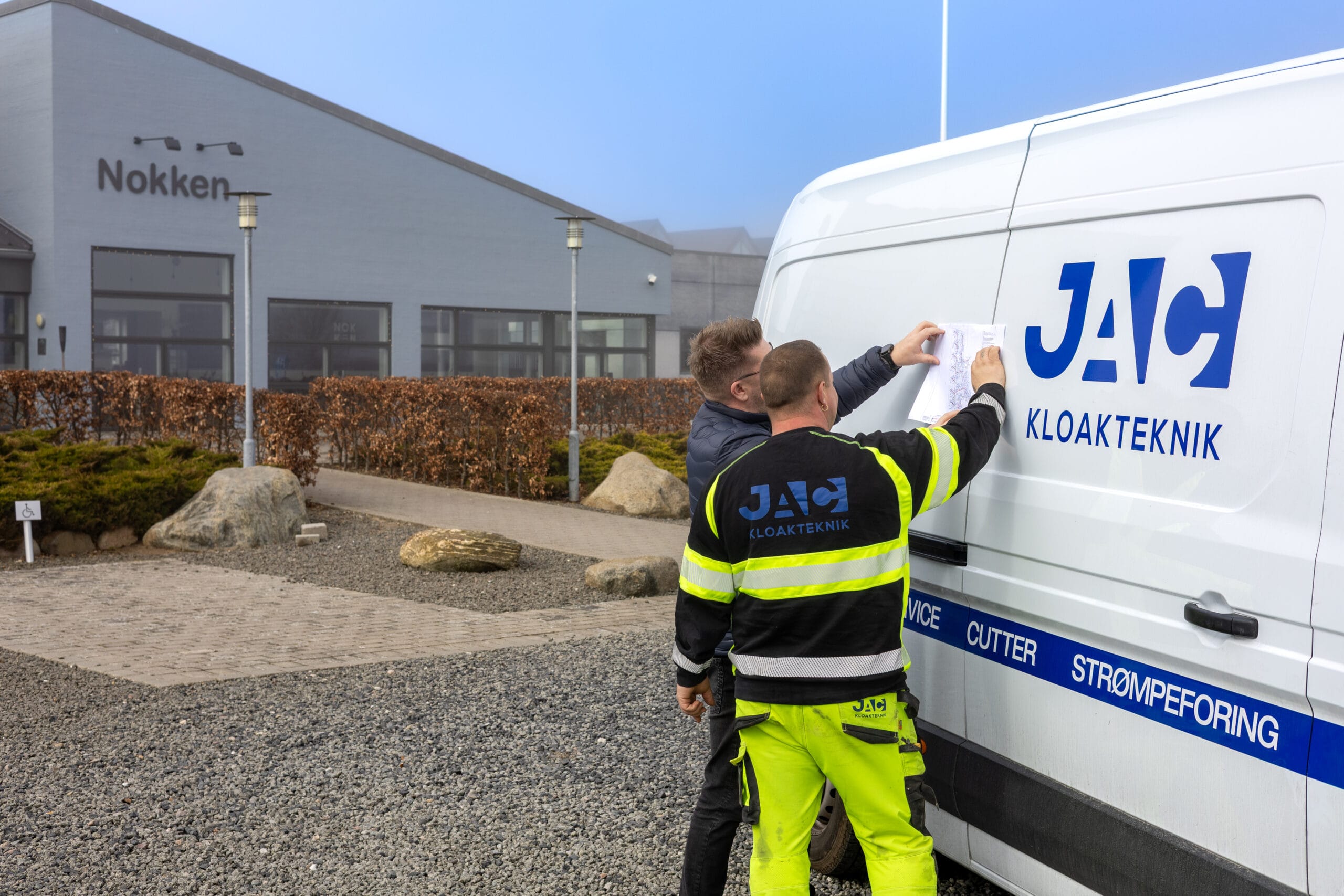 To medarbejdere fra JAC kloakteknik står ved en varevogn med teksten "JAC KLOAKTEKNIK" og "SERVICE CUTTER STRØMPEFORING" og gennemgår tegninger til faldstammerens. De er placeret foran bygningen med skiltet "Nokken" mens de arbejder udendørs i København.