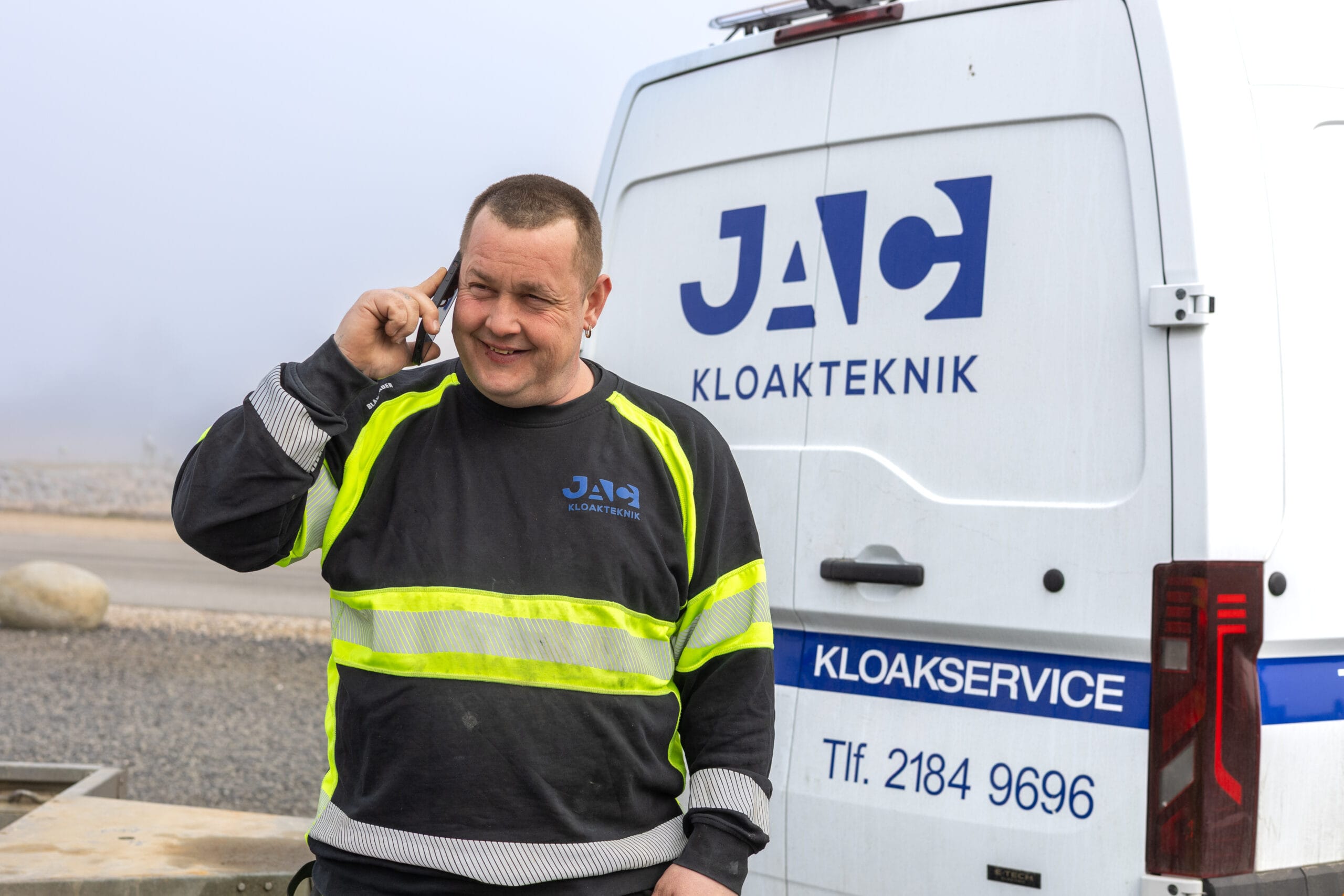 Medarbejder fra JAC kloakteknik står ved en varevogn med teksten "JAC KLOAKTEKNIK", "KLOAKSERVICE" og "Tlf. 2184 9696" og taler i telefon i forbindelse med faldstammerens. Han bærer arbejdstøj med refleksstriber og smiler foran bilen i København.