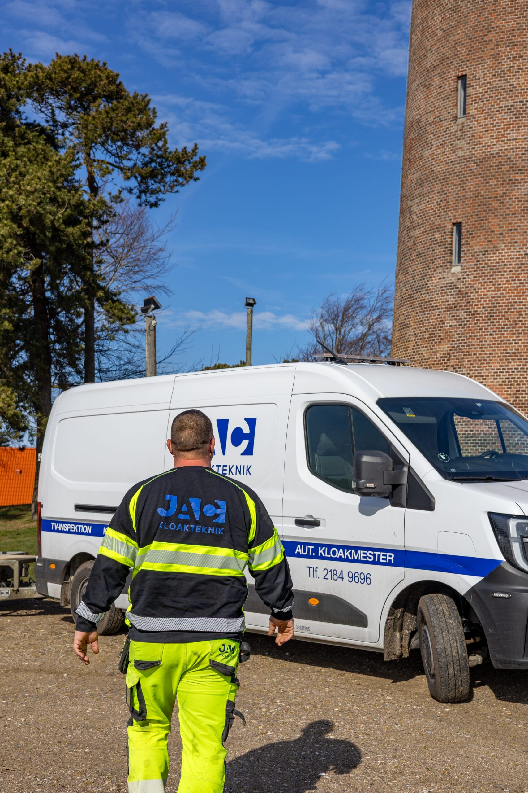 Medarbejder fra JAC kloakteknik går hen mod en varevogn med teksten "JAC KLOAKTEKNIK", "AUT. KLOAKMESTER" og "Tlf. 2184 9696" for at udføre faldstammerens. Han bærer arbejdstøj med refleksstriber og bevæger sig på en grusplads ved siden af bygninger i København.