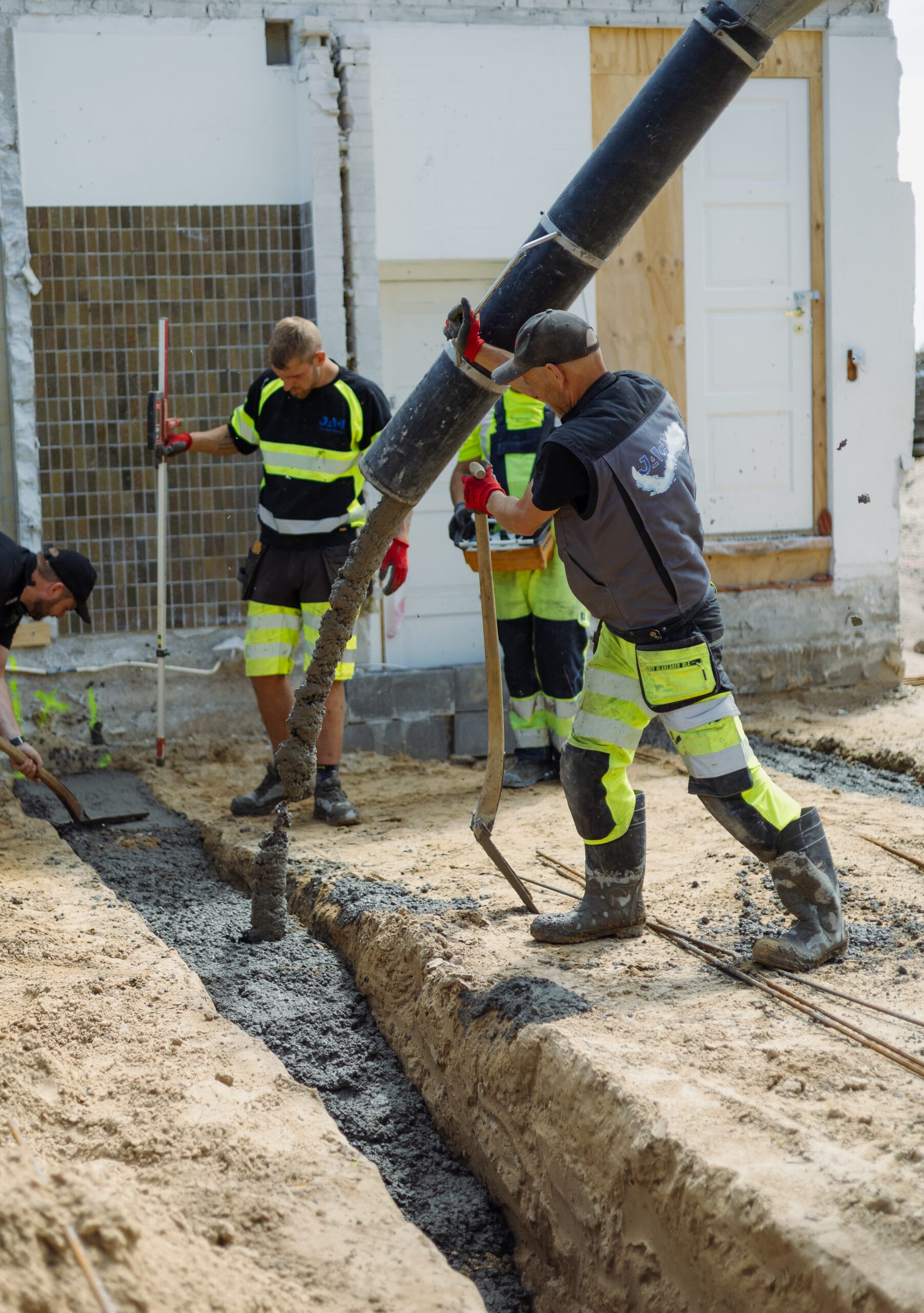 To kloakteknikere fra JAC Kloakservice lægger beton i en udgravet rende; en arbejder styrer en slange, der hælder våd beton ned i renden, mens en anden fordeler materialet med en skovl foran en bygning.