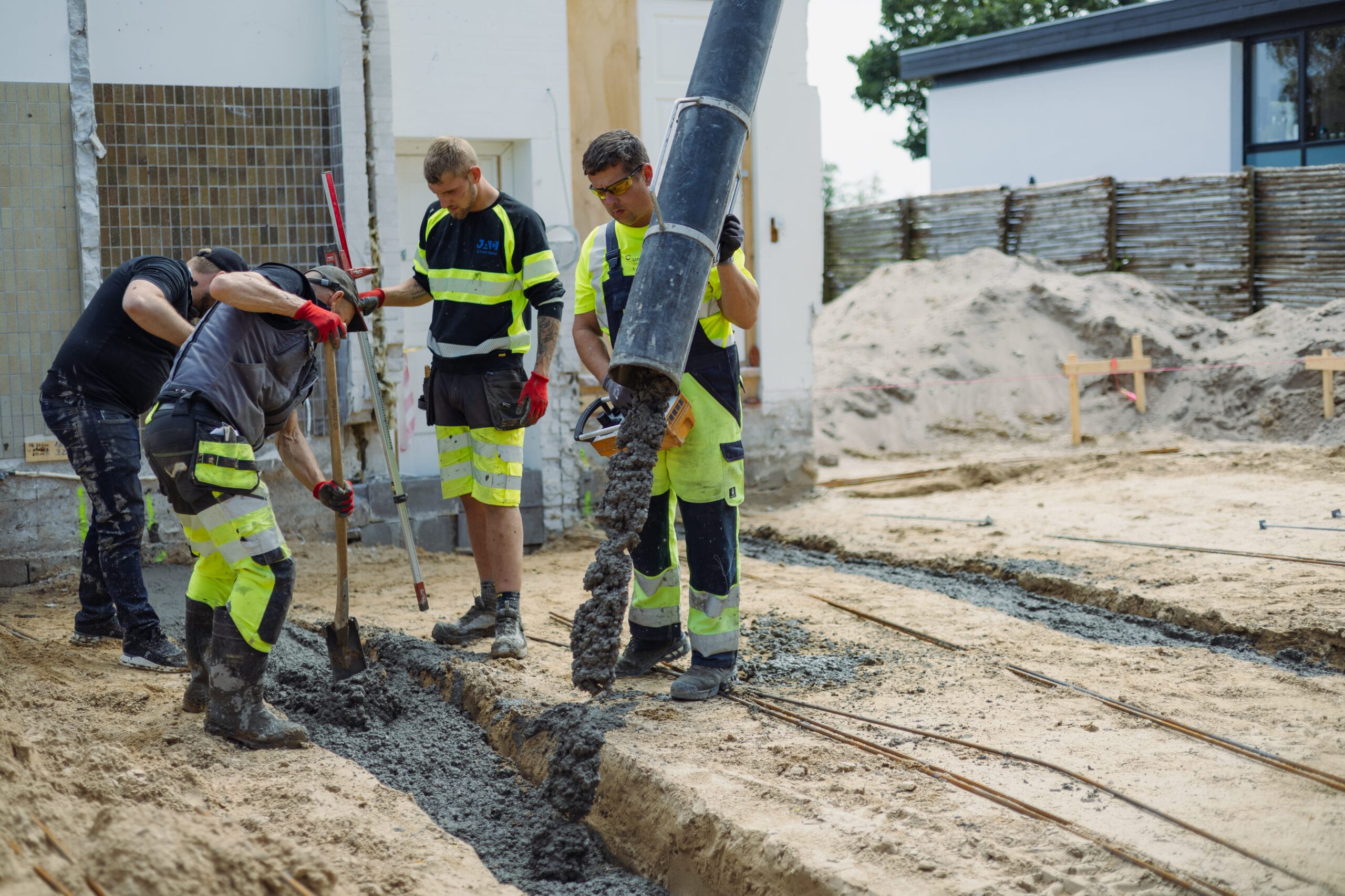 Flere kloakteknikere fra JAC Kloakservice udfører betonarbejde; en arbejder styrer en slange, der hælder våd beton ned i en udgravet rende, mens kollegaer fordeler og justerer betonen med værktøj på en byggeplads ved en bygning.