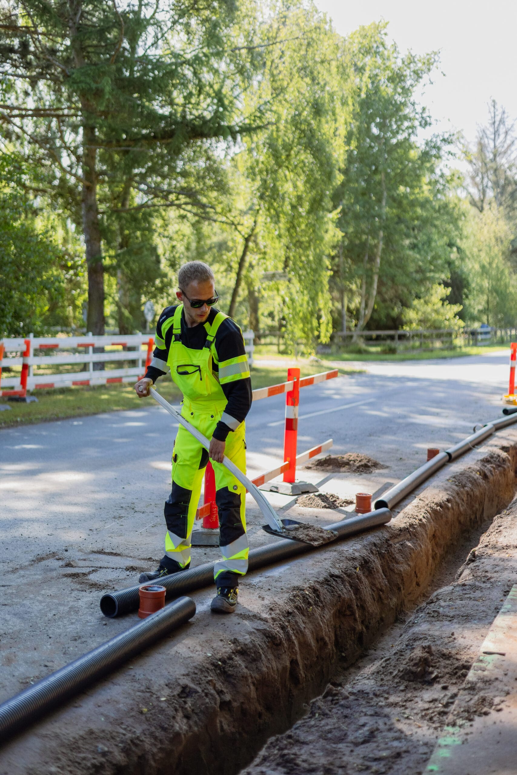 Kloaktekniker fra JAC Kloakservice arbejder på dræn i Rørvig; han står ved en udgravet rende langs vejen og fordeler jord med en skovl omkring nedlagte rør, med vejspærring og træer i baggrunden.