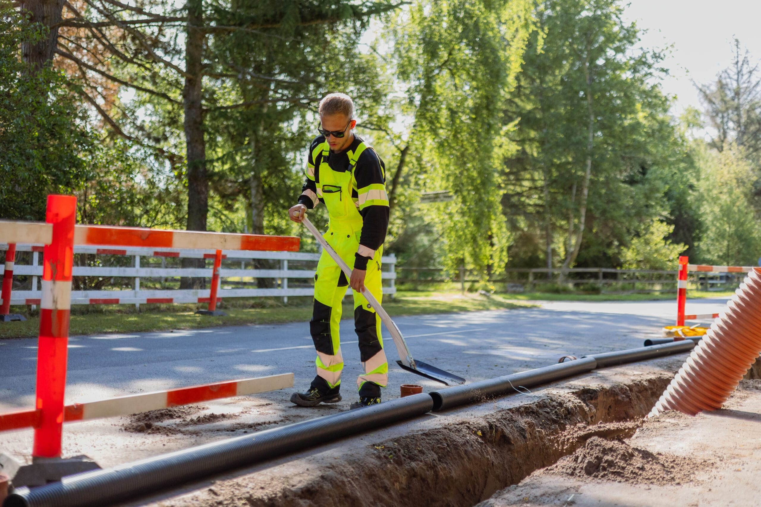 Kloaktekniker fra JAC Kloakservice arbejder på dræn i Rørvig; han står ved en udgravet rende langs vejen og bruger en skovl til at justere jord omkring et nedlagt rør, iført fluorescerende arbejdstøj, mens afspærring og træer ses i baggrunden.