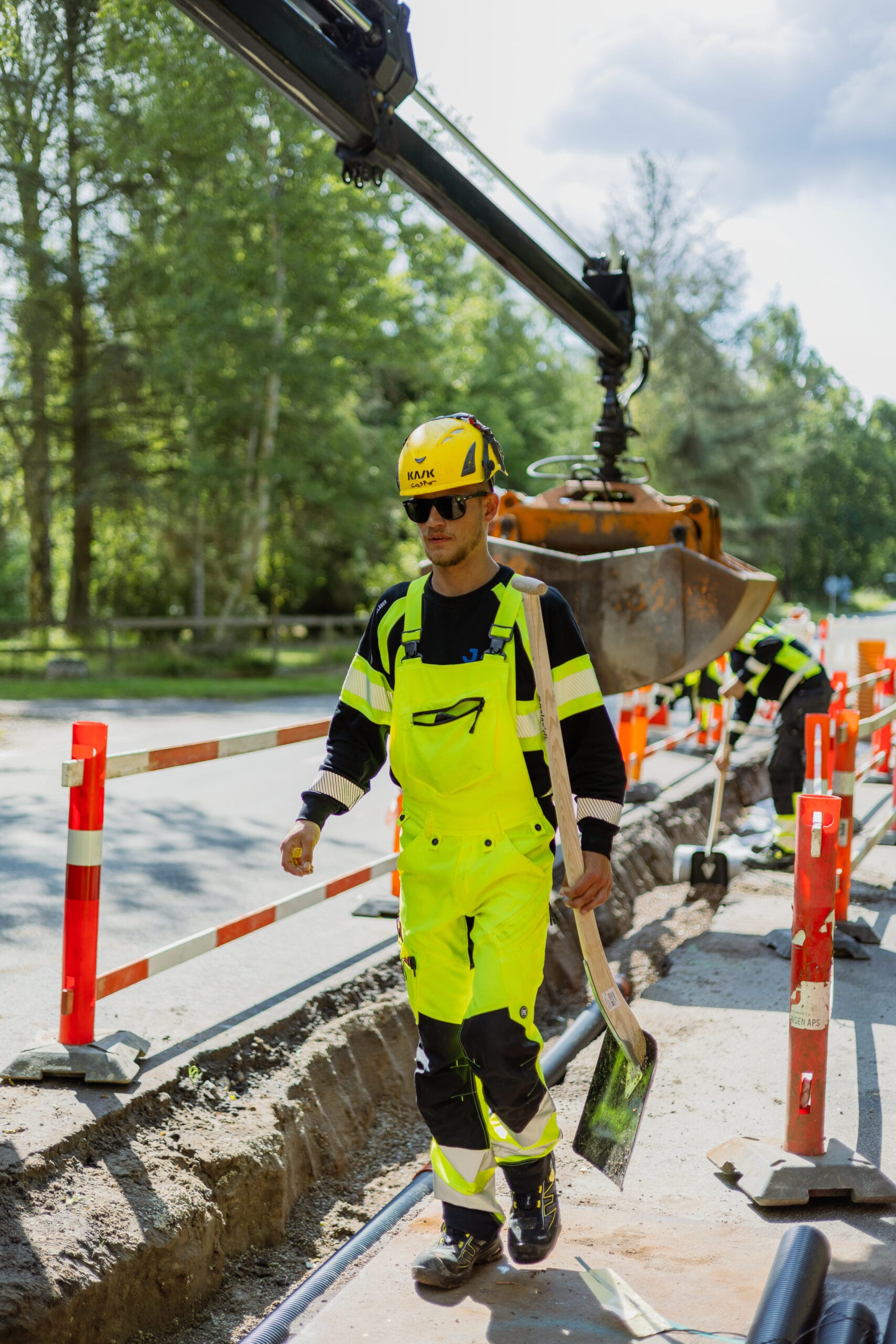 Kloaktekniker fra JAC Kloakservice arbejder på dræn i Rørvig; han går langs en udgravet rende med en skovl, iført gul sikkerhedshjelm og fluorescerende arbejdstøj, mens en gravemaskine og kollegaer arbejder i baggrunden ved vejarbejde.