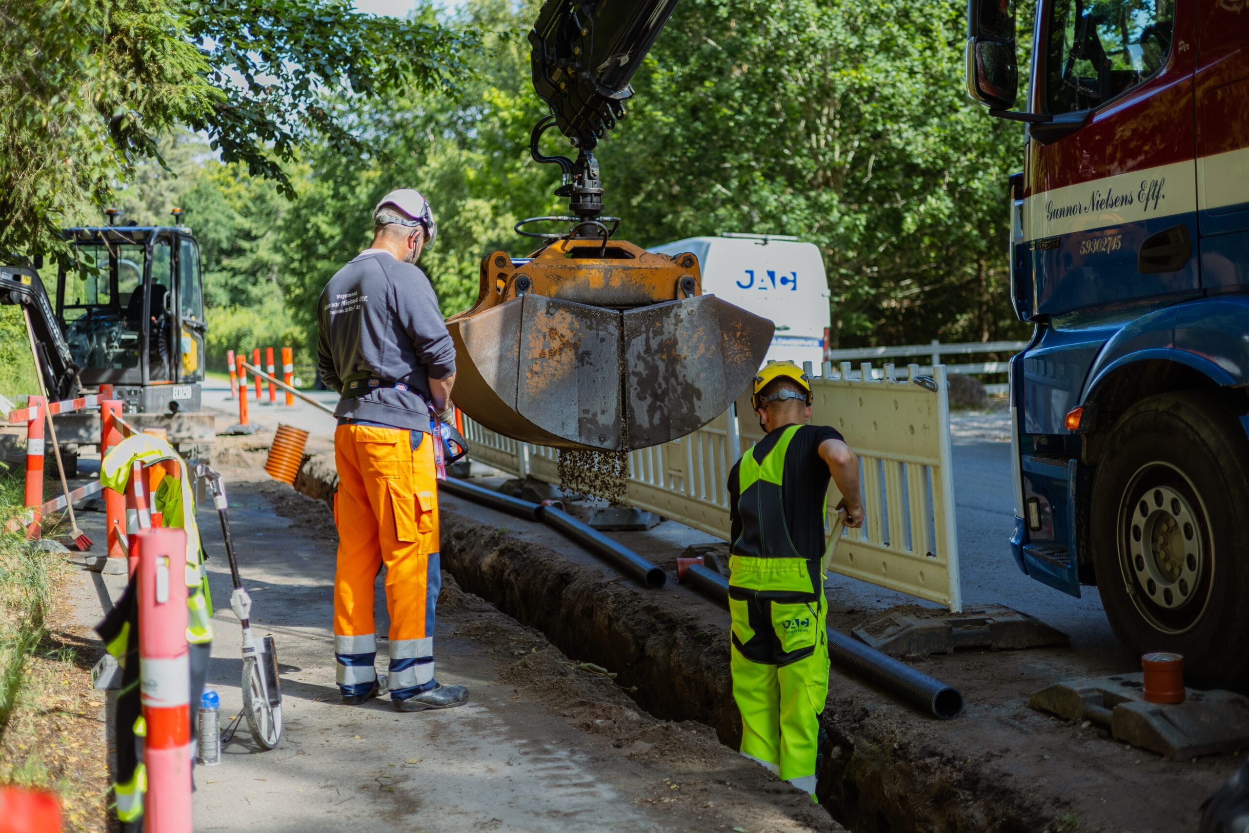 Kloaktekniker fra JAC Kloakservice arbejder på dræn i Rørvig; to arbejdere står ved en udgravet rende langs vejen, mens en gravemaskine sænker grus ned omkring kloakrør, med afspærring og servicekøretøj i baggrunden.