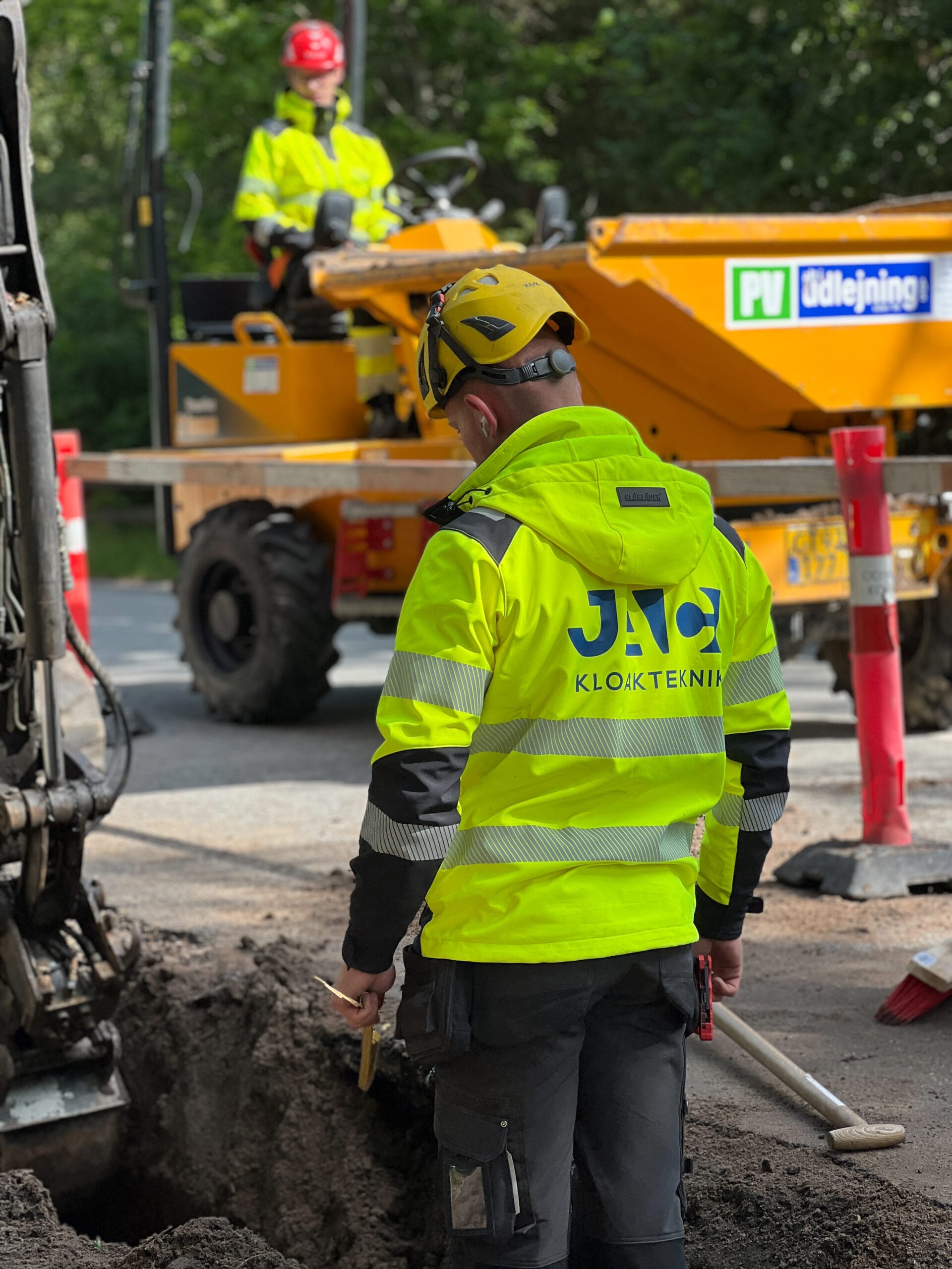 Kloaktekniker fra JAC Kloakservice arbejder på dræn i Rørvig; set bagfra står han ved en udgravet rende med værktøj i hånden, iført gul sikkerhedsjakke og hjelm, mens entreprenørmaskine og kollega ses i baggrunden ved vejarbejde.