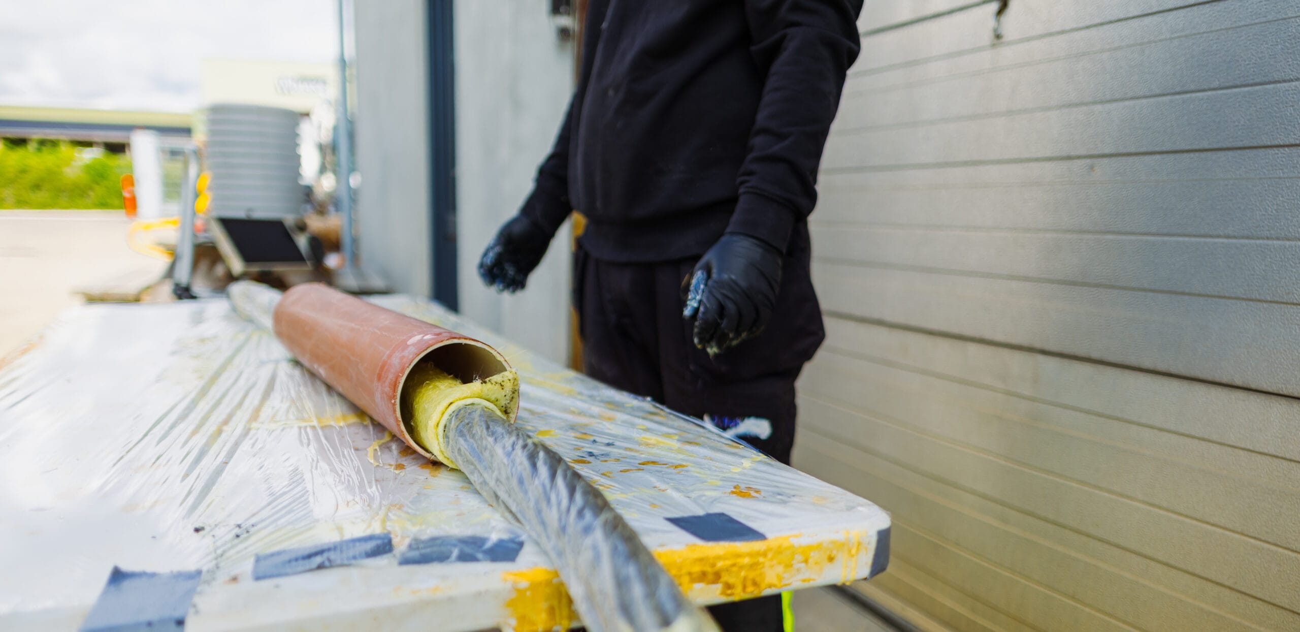 En JAC kloaktekniker står ved et arbejdsbord og arbejder med strømpeforing. En gul imprægneret foringsstrømpe føres ind i et rør, som ligger på bordet dækket af plast og harpiksrester.